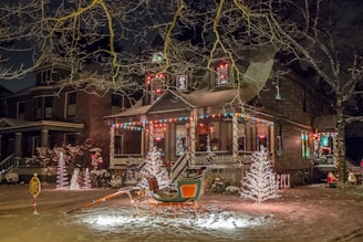A festive house adorned with colorful holiday lights and decorations, including illuminated trees and a sleigh in the front yard. Snow covers the ground, adding to the wintery and cheerful atmosphere.
