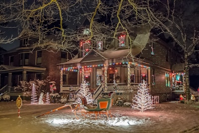 A festive house adorned with colorful holiday lights and decorations, including illuminated trees and a sleigh in the front yard. Snow covers the ground, adding to the wintery and cheerful atmosphere.