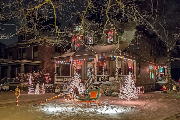 A festive house adorned with colorful holiday lights and decorations, including illuminated trees and a sleigh in the front yard. Snow covers the ground, adding to the wintery and cheerful atmosphere.