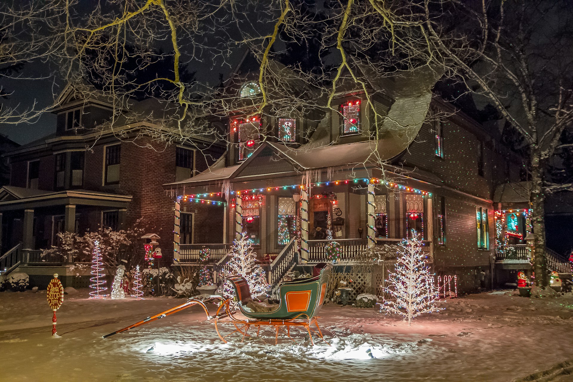 A festive house adorned with colorful holiday lights and decorations, including illuminated trees and a sleigh in the front yard. Snow covers the ground, adding to the wintery and cheerful atmosphere.