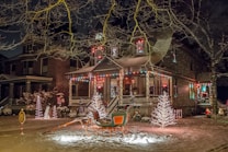 A festive house adorned with colorful holiday lights and decorations, including illuminated trees and a sleigh in the front yard. Snow covers the ground, adding to the wintery and cheerful atmosphere.