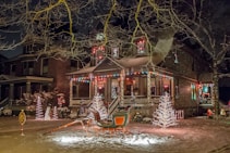 A festive house adorned with colorful holiday lights and decorations, including illuminated trees and a sleigh in the front yard. Snow covers the ground, adding to the wintery and cheerful atmosphere.
