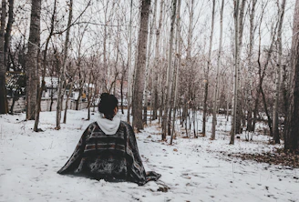 man in green and black robe sitting on snow covered ground surrounded by bare trees during
