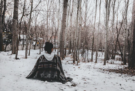 man in green and black robe sitting on snow covered ground surrounded by bare trees during