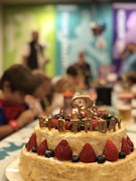 A festive scene featuring a layered cake adorned with large strawberries and blueberries. The cake has the word 'BIRTHDAY' spelled out on top in decorative letters. Children are gathered around a table in the background, appearing to be at a party.