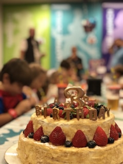A festive scene featuring a layered cake adorned with large strawberries and blueberries. The cake has the word 'BIRTHDAY' spelled out on top in decorative letters. Children are gathered around a table in the background, appearing to be at a party.