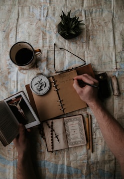 A person's hands are visible; one hand is writing in an open brown notebook with a black pen, and the other is holding a small book depicting a crucifixion. The notebook lies on a spread-out map. Nearby objects include a cup of coffee, a pair of glasses, a small round metal object with a symbol, a green succulent plant, a knife, a rosary, and a second small open book.
