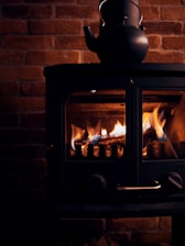 A black kettle sits on top of a dark, compact wood-burning stove with a glass front. The glow of the flames inside the stove illuminates the bricks of the surrounding wall, casting a warm, cozy light.