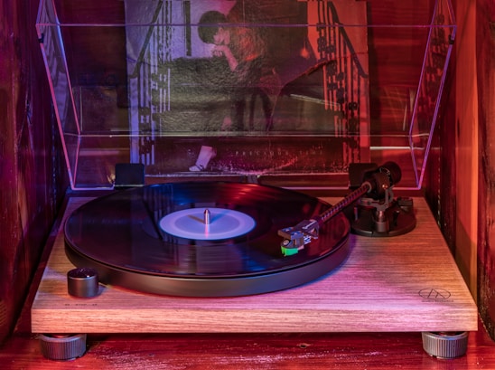 A turntable with a vinyl record is displayed on a wooden surface, partially enclosed by a clear protective case. Behind the turntable, there's a large poster with a person leaning against a patterned background, lit by moody, colored lighting.