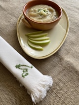 A bowl of creamy soup is placed on an oval plate alongside several slices of green apple. The setting includes a tablecloth with a natural texture and a neatly folded white napkin embroidered with green leaves.