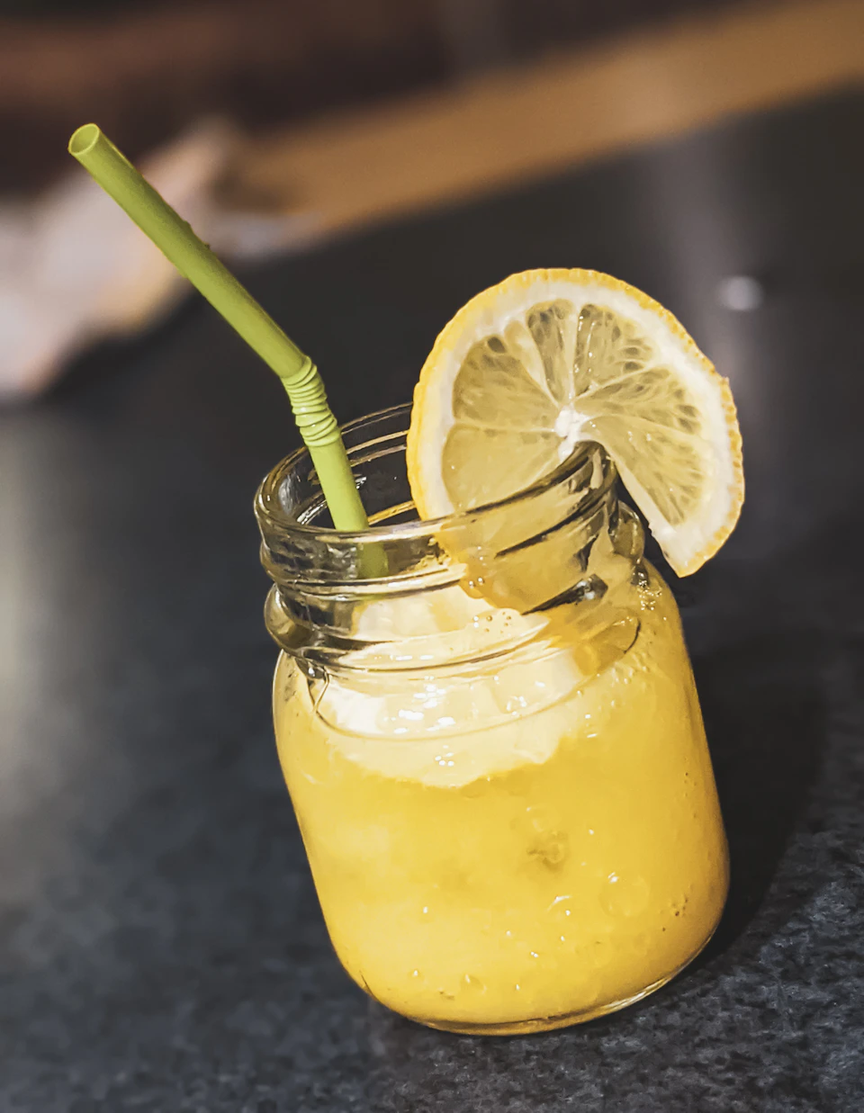 The image shows a tall glass jar filled with a vibrant yellow liquid, likely lemonade or another type of sweetened beverage. The jar is placed on a black surface and has a green straw protruding from it. A slice of lemon rests atop the drink in the jar, adding a pop of color to the scene.