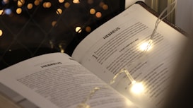 An open book titled 'Hebrews' is illuminated by string lights, with a background of warm, blurry bokeh lights.