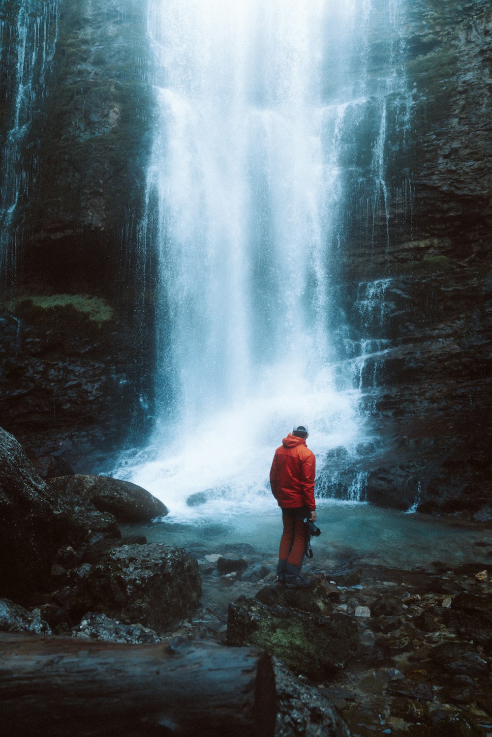man in orange jacket standing on rock near waterfalls during daytime