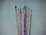 A collection of nine paintbrushes with colorful, decorated handles arranged vertically against a textured blue background. The handles feature patterns in shades of orange, blue, pink, green, and silver. The brush sizes vary from fine to medium.