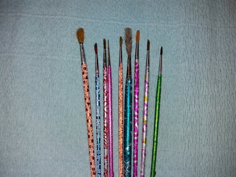 A collection of nine paintbrushes with colorful, decorated handles arranged vertically against a textured blue background. The handles feature patterns in shades of orange, blue, pink, green, and silver. The brush sizes vary from fine to medium.