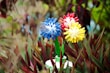 Artificial flowers made from colorful cylindrical elements arranged in a spherical design are placed amidst spiky green and reddish-brown foliage. The flowers come in blue, red, and yellow with stems in a flowerpot.