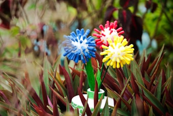 Artificial flowers made from colorful cylindrical elements arranged in a spherical design are placed amidst spiky green and reddish-brown foliage. The flowers come in blue, red, and yellow with stems in a flowerpot.