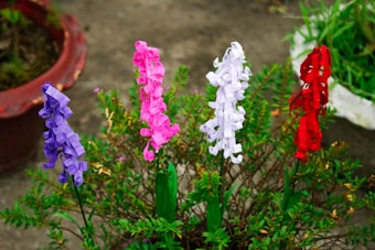 There are several paper flowers on display, each crafted in different colors including purple, pink, white, and red. They are placed among green foliage and small plants in a garden setting. A clay pot partially filled with greenery is visible in the background.