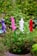 Colorful artificial flowers in purple, pink, white, and red are arranged in a lush green potted plant against a blurred background of outdoor plants.