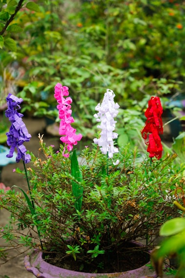 Colorful artificial flowers in purple, pink, white, and red are arranged in a lush green potted plant against a blurred background of outdoor plants.