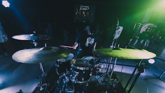 A person playing a drum set in a dimly lit room with vibrant green and blue lighting. Several drums and cymbals are visible, as well as microphones and music equipment. In the background, there is a projected image on a screen with blurred figures and text.