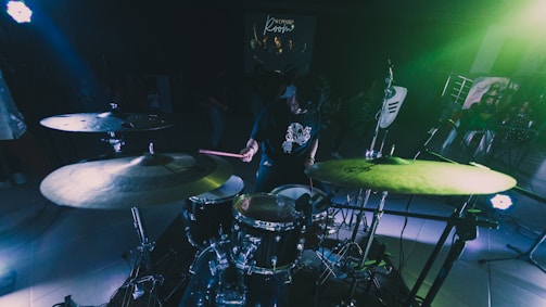A person playing a drum set in a dimly lit room with vibrant green and blue lighting. Several drums and cymbals are visible, as well as microphones and music equipment. In the background, there is a projected image on a screen with blurred figures and text.