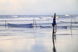A serene scene at a beach with gentle waves rolling against the shore. Wooden stakes are set up in the shallow water, supporting blue fishing nets. The nets are slightly elevated above the water surface, awaiting the tide.