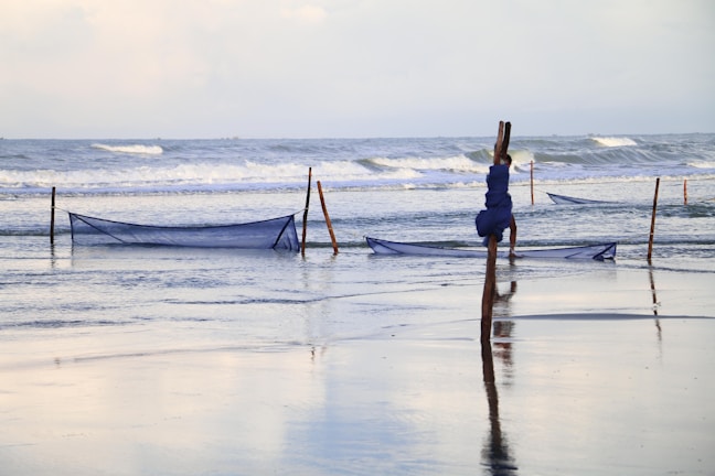 A serene scene at a beach with gentle waves rolling against the shore. Wooden stakes are set up in the shallow water, supporting blue fishing nets. The nets are slightly elevated above the water surface, awaiting the tide.