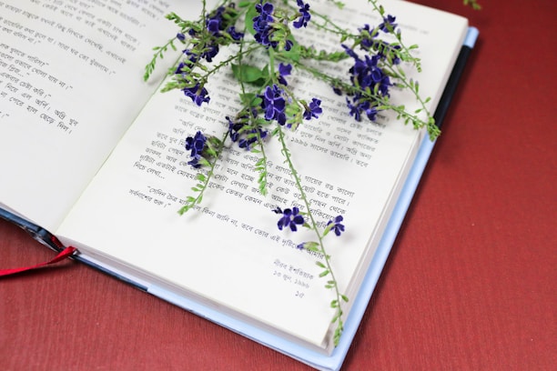 An open book with text in a South Asian script, possibly Bengali, lies on a flat surface. A sprig of delicate purple flowers rests across the pages, adding a decorative and contemplative element. The background is a solid, warm red, enhancing the contrast with the book and flowers.