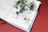 An open book with text in a South Asian script, possibly Bengali, lies on a flat surface. A sprig of delicate purple flowers rests across the pages, adding a decorative and contemplative element. The background is a solid, warm red, enhancing the contrast with the book and flowers.