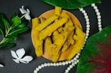 Several pieces of turmeric root arranged on a round wooden platform, surrounded by green leaves and white flowers, with a string of white pearls beside them. The background is a soft, dark surface enhancing the warm yellow tones of the turmeric and the lush green of the leaves.