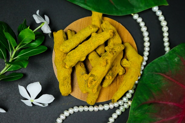 Several pieces of turmeric root arranged on a round wooden platform, surrounded by green leaves and white flowers, with a string of white pearls beside them. The background is a soft, dark surface enhancing the warm yellow tones of the turmeric and the lush green of the leaves.