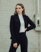 A person in a business suit stands against a light gray brick wall. They are wearing a dark blazer and pants over a white turtleneck, with their hand on their hip in a confident pose. An electrical box and conduit can be seen in the background.