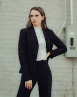 A person in a business suit stands against a light gray brick wall. They are wearing a dark blazer and pants over a white turtleneck, with their hand on their hip in a confident pose. An electrical box and conduit can be seen in the background.
