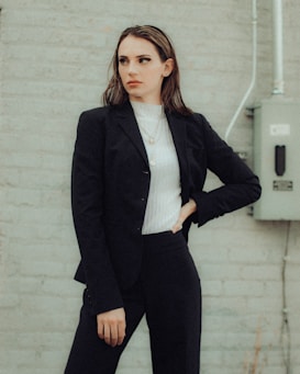 A person in a business suit stands against a light gray brick wall. They are wearing a dark blazer and pants over a white turtleneck, with their hand on their hip in a confident pose. An electrical box and conduit can be seen in the background.