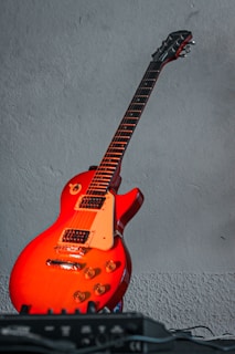 A vibrant red electric guitar with a classic Les Paul shape is leaning against a textured grey wall. The guitar features a mahogany body with a glossy finish and white pickguard, showcasing multiple control knobs and a pickup selector switch. In the foreground, an electronic device is partially visible, possibly an amplifier or effects pedal.