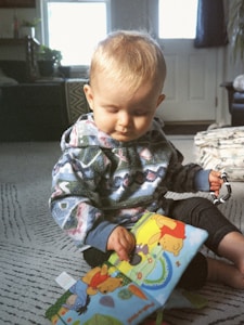 A young child with blonde hair is sitting on a patterned carpet, intently looking at a colorful book. The child is wearing a patterned sweater and holding a small toy in their hand. The background features a sofa, a window with bright light streaming through, and a door.