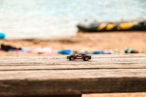 A small toy car with red wheels and a black body sits on a wooden surface. The background shows a blurred sandy beach and the calm waters of a lake, suggesting a serene lakeside setting.