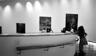 A group of people are observing large paintings displayed on the walls of an art gallery. The gallery features a modern design with curving walkways and bright, diffused lighting from above. The paintings appear to be detailed, and the visitors are facing different artworks, engrossed in contemplation.