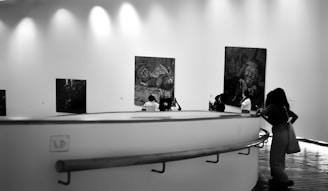 A group of people are observing large paintings displayed on the walls of an art gallery. The gallery features a modern design with curving walkways and bright, diffused lighting from above. The paintings appear to be detailed, and the visitors are facing different artworks, engrossed in contemplation.