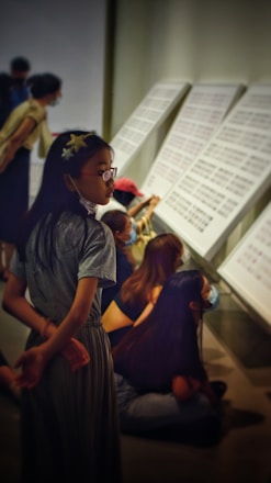 A group of people, mostly young, are engaged in observing large panels with text or patterns on them. The environment suggests a gallery or exhibition space. All are wearing casual clothing, and some are sitting on the floor while others stand or lean forward, indicating a focused attention on the display.