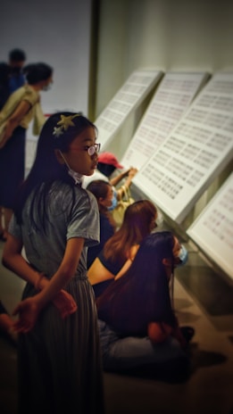 A group of people, mostly young, are engaged in observing large panels with text or patterns on them. The environment suggests a gallery or exhibition space. All are wearing casual clothing, and some are sitting on the floor while others stand or lean forward, indicating a focused attention on the display.