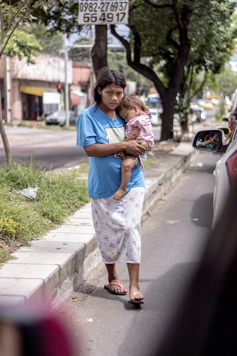 woman in blue t-shirt and white floral skirt carrying baby in white dress