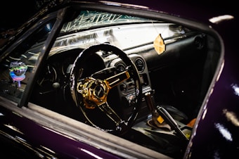 A close-up view of a car interior featuring a shiny, gold-colored steering wheel and a manual gear shift. The dashboard includes various gauges and a hanging air freshener is visible.