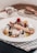 A plate with scoops of ice cream decorated with chocolate sprinkles, a raspberry on top, and surrounded by colorful fruits like strawberries and peaches. A biscuit stick is laid across the ice cream. In the background, a cup of coffee and a set of wooden cutlery on a coaster are placed on a patterned tablecloth.