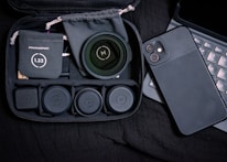 A collection of phone camera accessories is neatly arranged. A black phone case and multiple camera lens attachments are visible. Each lens is in its individual compartment inside a black zippered case. A digital device with a keyboard is partially visible.