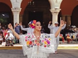 A person dressed in traditional clothing featuring intricate floral embroidery and lace details, with colorful flowers in their hair. They are participating in a dance, surrounded by a group of people dressed in white clothing, some of whom are clapping along. The background shows architectural elements with arches.