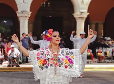 A person dressed in traditional clothing featuring intricate floral embroidery and lace details, with colorful flowers in their hair. They are participating in a dance, surrounded by a group of people dressed in white clothing, some of whom are clapping along. The background shows architectural elements with arches.