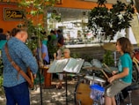 A young person is playing a drum set outdoors next to an adult playing a stringed instrument, possibly a guitar. They are surrounded by trees and plants, and several people are seated at tables in the background. An older woman holds a fan while observing the scene. The setting appears to be a café or restaurant with an orange exterior and a relaxed atmosphere.