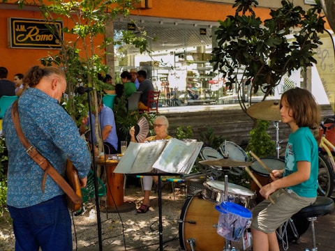 A young person is playing a drum set outdoors next to an adult playing a stringed instrument, possibly a guitar. They are surrounded by trees and plants, and several people are seated at tables in the background. An older woman holds a fan while observing the scene. The setting appears to be a café or restaurant with an orange exterior and a relaxed atmosphere.